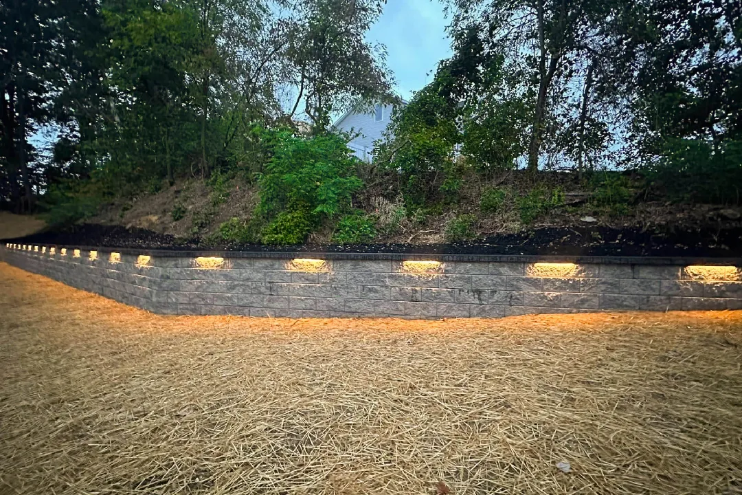 Gray block retaining wall with warm recessed lighting below a tree-lined slope