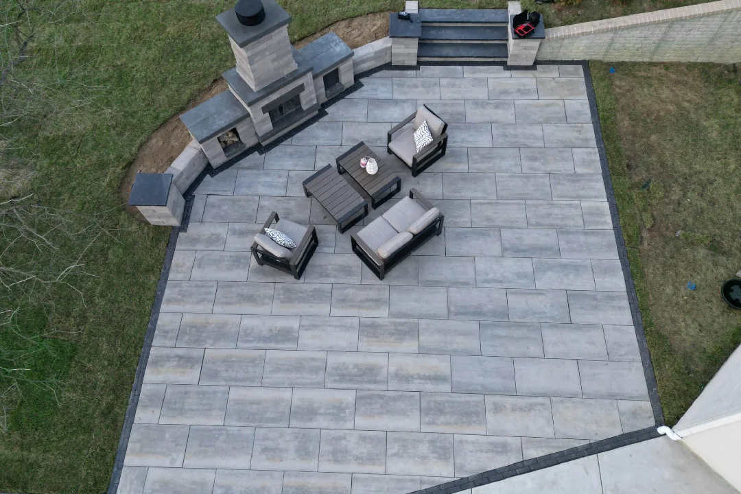 Finished gray paver patio with an outdoor fireplace, cushioned seating, and steps through a block wall